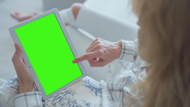 Young Woman in white jeans sitting on couch uses Tablet PC with pre-keyed green screen. Few types of gestures - scrolling up and down, tapping, zoom in and out. Perfect for screen compositing