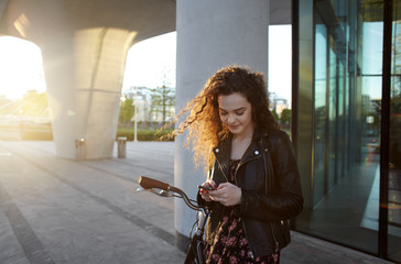 Fototapeta na wymiar Beautiful teenage girl with curly hair typing text message to her boyfriend on electronic device during bike ride on vintage bicycle, standing in urban setting, smiling as she reads sms. Flare sun