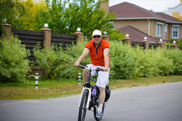 A mature man  in shorts, a T-shirt and a cap rides a bicycle around the cottage town. The concept of a healthy lifestyle after 50 years