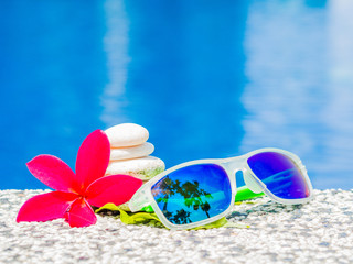 Sunglasses with red frangipani (plumeria) flowers and stack of stone at the side of swimming pool. Vacation, beach, summer travel concept