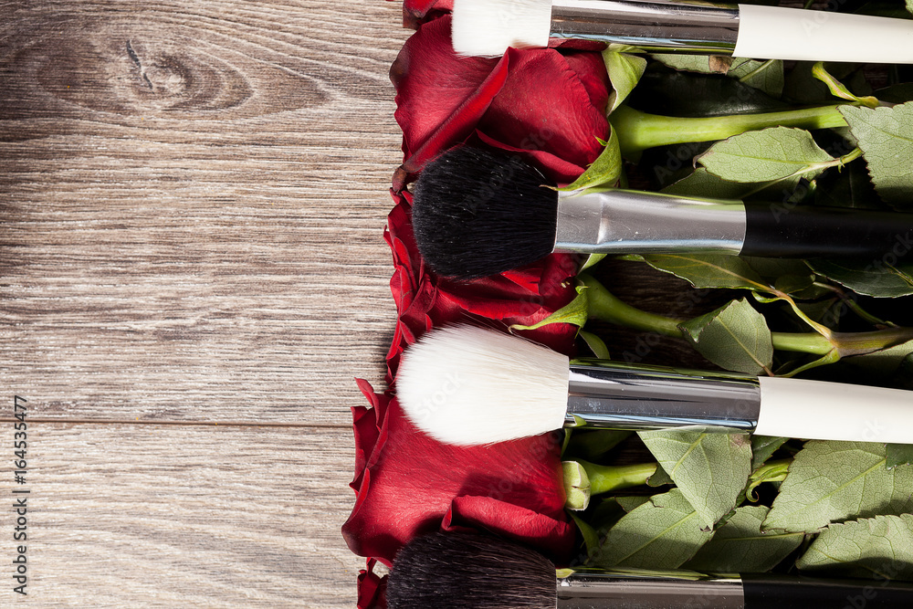 Wall mural Conceptual image of make-up brushes next to roses on wooden background