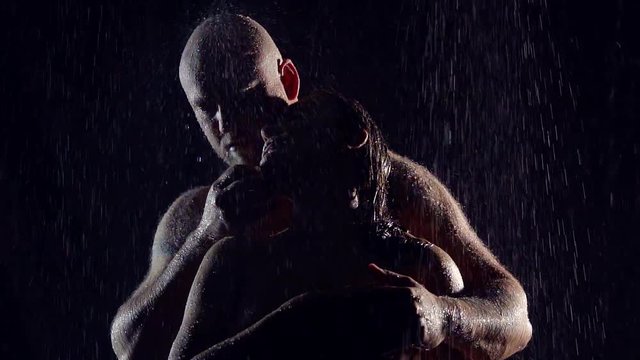 silhouette in the darkness under the big jets of water from the rain a couple sporty man with bald naked cuddles up to the woman and caresses her hands.