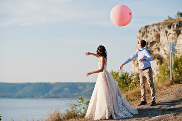 Gorgeous bride and groom having great time standing on the precipice with a view of a lake with balloons in their hands on their wedding day.