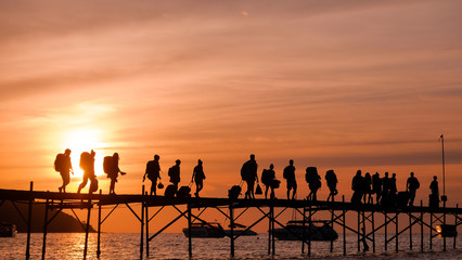 Silhouettes of the backpackers walking along the wooden pier to the ferry on beautiful sunset time. Tourists with bags walk along the Bang Rack pier to the ferry to Phangan island