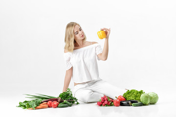 Beautiful blond woman in white clothes and lots of fresh vegetables on white background. Girl holds bell pepper