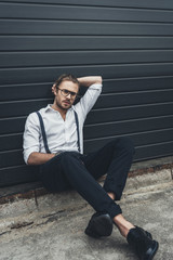 Handsome pensive stylish man in eyeglasses sitting with hand behind head on pavement and looking away
