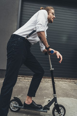 Side view of stylish young man wearing eyeglasses and suspenders riding scooter