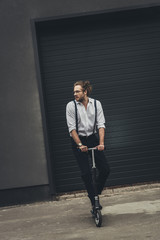 Stylish young man in eyeglasses and suspenders standing on scooter and looking away