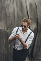 Portrait of handsome stylish bearded man in sunglasses and suspenders lighting cigarette with lighter