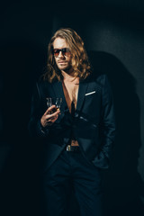 Handsome bearded long haired man in sunglasses and stylish suit holding glass of whiskey on black