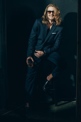 Handsome young man in suit and sunglasses holding glass of whisky and smiling at camera