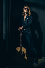 Handsome long haired man in sunglasses and stylish suit posing with acoustic guitar
