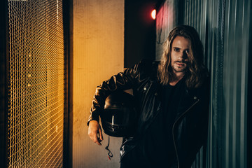 Stylish bearded man in leather jacket holding helmet and looking at camera