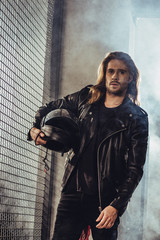 Stylish long haired bearded man in leather jacket holding helmet and looking at camera