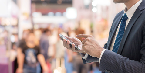 Businessman using smartphone Communicate at trade shows exhibition hall with full of people walking blurred background. - obrazy, fototapety, plakaty