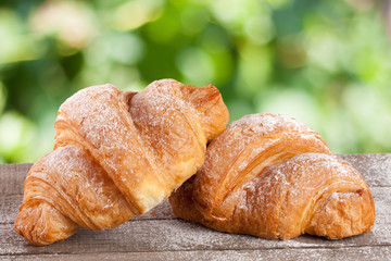 two croissant sprinkled with powdered sugar on a wooden board with a blurry garden background