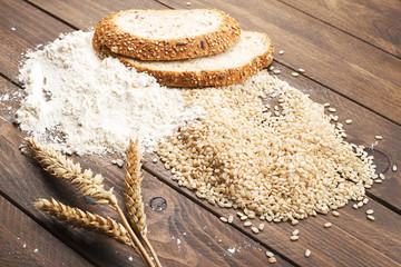 Flour and rice next to bread and wheat flower on wooden table.