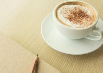 Hot latte in white cup, pencil and notebook on wood table.