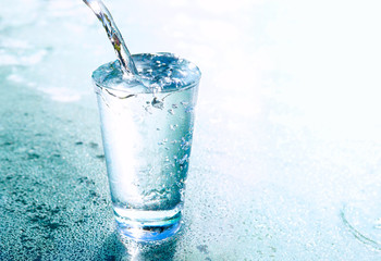 Een stroom helder transparant koud water wordt gegoten in een glazen beker op blauwe achtergrond met mooie verlichting close-up. Water gloeit in een glas.