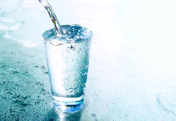 Foto op Plexiglas Water Een stroom helder transparant koud water wordt gegoten in een glazen beker op blauwe achtergrond met mooie verlichting close-up. Water gloeit in een glas.