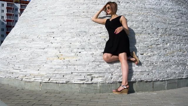 Stylish fashionable young female model in sunglasses in a black dress possing outdoor.