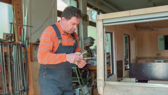 A middle-aged woodworker is using two types of wrenches to adjust the car mirror on a vintage car that he is designing in his workshop at home. 