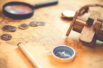 Compass on vintage world map with coins, pen, wrist watch, plane and Magnifying glass for vacation and travel concept, selective focus.
