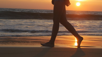 Legs of young woman going along ocean beach during sunrise. Female feet walking barefoot on sea shore at sunset. Girl stepping in shallow water at shoreline. Summer vacation concept. Close up