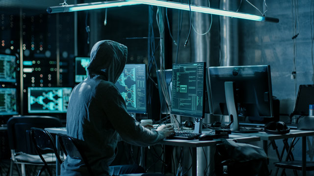 Hooded Hacker Using His Comuter with Different Information to Break into Corporate Data Servers and Infects them with Virus. His Hideout Place has Dark Atmosphere, Multiple Displays.