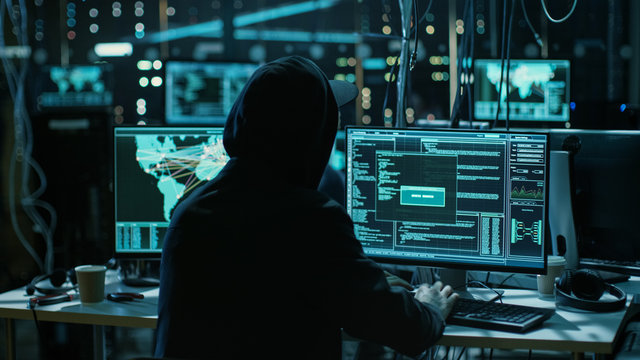 Teenage Hacker Working with His Computer Infecting Servers and Infrastructure with Malware. His Hideout is Dark and Has Multiple displays.