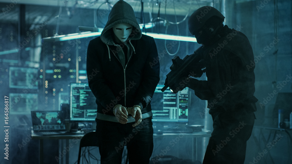 Poster Masked Hacker Wearing Handcuffs is Guarded by Fully Armed Special Forces Soldier.  They're in Hacker's Hideout Basement with Multiple Operating Displays.