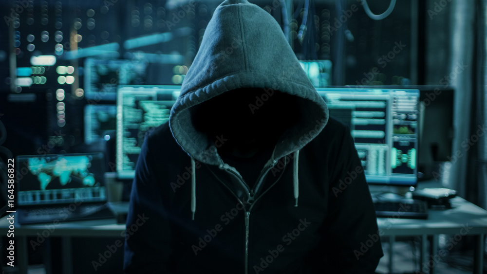 Aufkleber Dangerous Internationally Wanted Hacker Wth Hided Face Looking into the Camera. In the Background His Dark Operating Room with Multiple Displays and Cables.