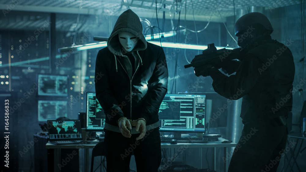 Poster Masked Hacker Wearing Handcuffs is Guarded by Fully Armed Special Forces Soldier.  They're in Hacker's Hideout Basement with Multiple Operating Displays.