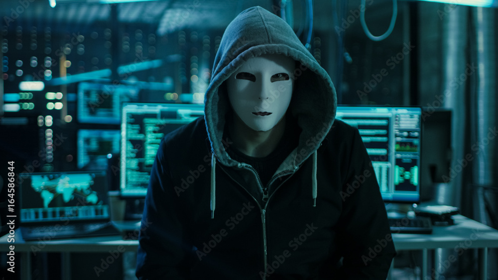 Poster Dangerous Internationally Wanted Hacker with Covered Face Looking into the Camera. In the Background His Operating Room with Multiple Displays and Cables.