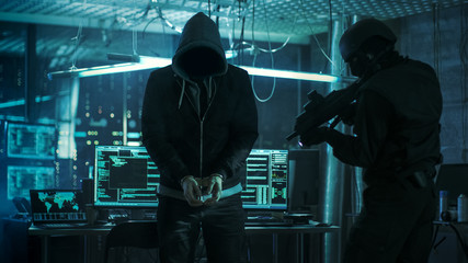 Handcuffed Masked Hacker is Standing and Guarded by Fully Armed Special Forces Soldier. They're in Hacker's Hideout Basement with Multiple Operating Displays.