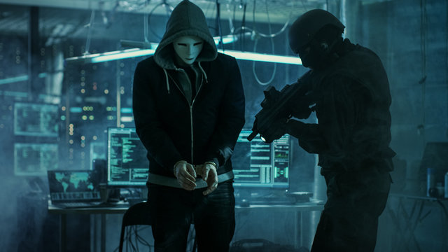 Masked Hacker Wearing Handcuffs is Guarded by Fully Armed Special Forces Soldier.  They're in Hacker's Hideout Basement with Multiple Operating Displays.