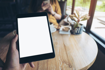 Mock up image of a hand holding black tablet pc with white blank screen on wooden table with woman using mobile phone in background in modern cafe