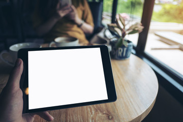 Mock up image of a hand holding black tablet pc with white blank screen on wooden table with woman using mobile phone in background in modern cafe