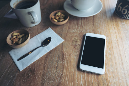 Mockup image of white mobile phone with blank black screen , coffee cups and snacks on vintage wooden table in cafe