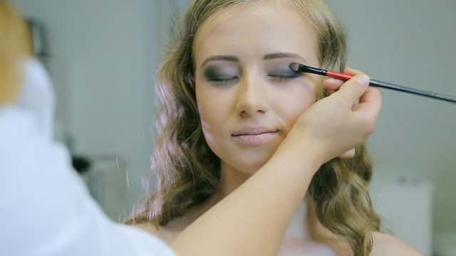 Professional makeup artist applying tonal powder to long curl hair blond models with blue eyelid for doing eye shadow. Front closeup view.