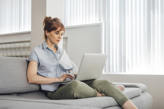 Beautiful Caucasian Woman Sitting On Couch At Home And Typing On Laptop.