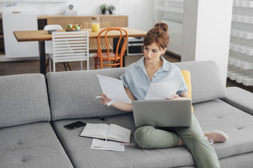 Beautiful ginger Caucasian woman holding laptop in her lap and reading from papers and sitting at home.