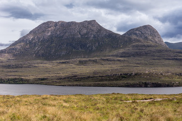 Assynt Peninsula, Scotland - June 7, 2012: Brown Mountain range along Loch Lurgainn under silver sky with gray clouds. Green vegetation on foothills and dry foreground.