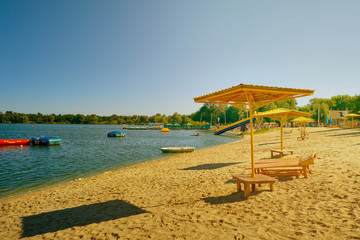 Summer sunny day. Beach, Wooden umbrella, sun loungers on the shore of a lake on the blue sky background.