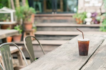 Cold black coffee drink with ice in plastic cup on wooden table with chairs in garden background 