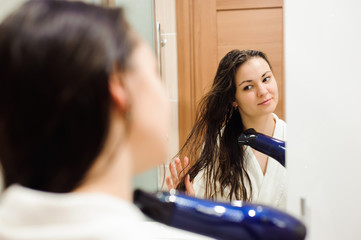 Beautiful young woman in bath towel is using a hair dryer and smiling while looking into the mirror in bathroom