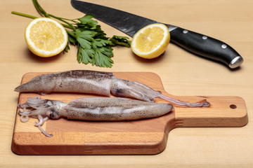Cooking fresh squids. Two raw cuttlefishes over a wooden cutting board in front of a sliced lemon, parsley and a knife, at kitchen