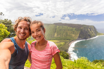 Selfie couple tourists at Waipio Valley lookout in Big Island, Hawaii. Happy smiling people taking self-portrait picture with camera phone. Woman and man taking photos on adventure nature travel.
