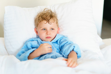 Little cute boy in blue pajamas with wet hair after bathing is lying on a huge snow-white pillow, wrapped in a blanket. He thoughtfully looks aside and positively grimaces.