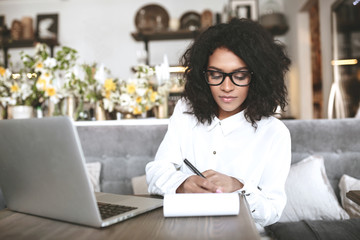 Young African American girl sitting in restaurant with laptop and notebook. Pretty girl in glasses working at cafe. Portrait of thoughtful lady with dark curly hair writing note in her notebook